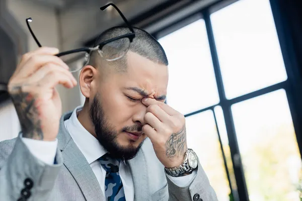 Erschöpfter asiatischer Geschäftsmann reibt sich die Augen, während er verschwommene Brillen im Büro hält — Stockfoto