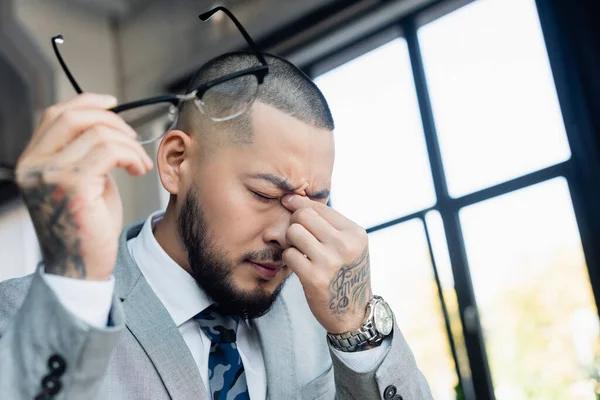 Exhausted asian businessman rubbing eyes while holding blurred eyeglasses in office — Stock Photo