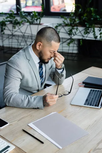 Tired asian businessman holding eyeglasses and rubbing eyes near laptop at workplace — Stock Photo