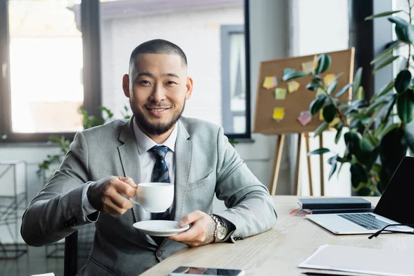 Feliz asiático hombre de negocios con taza de café mirando cámara mientras sentado en el lugar de trabajo - foto de stock