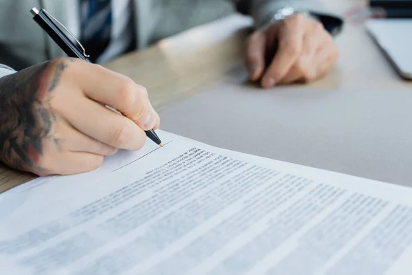 Partial view of tattooed businessman signing blurred contract in office — Stock Photo