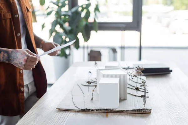 Teilansicht tätowierter Ingenieur mit Dokument in der Nähe verschwommener Hausmodelle auf dem Schreibtisch — Stockfoto