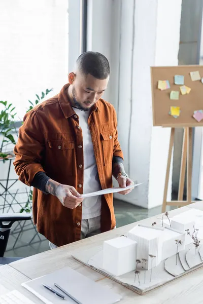 Asiatischer Architekt betrachtet Dokument, während er in der Nähe von Hausmodellen auf dem Schreibtisch steht — Stockfoto