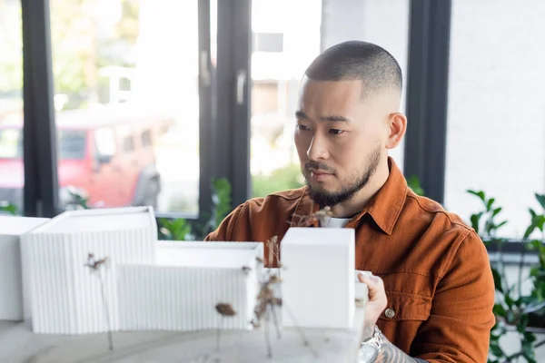 Bearded asian architect looking at blurred construction project with house models — Stock Photo