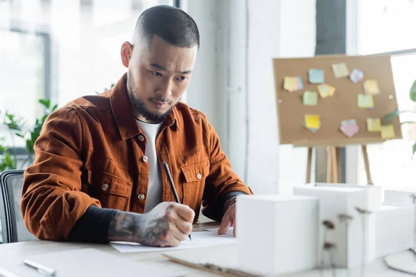 Tattooed asian architect looking at blurred house models while drawing blueprint in office — Stock Photo