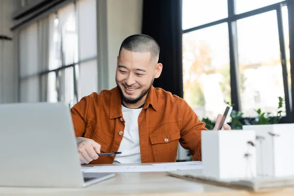 Smiling asian architect holding smartphone while working with blueprints near blurred laptop — Stock Photo