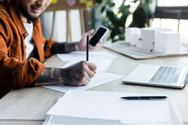 Cropped view of blurred architect with mobile phone drawing blueprint while working in office — Stock Photo