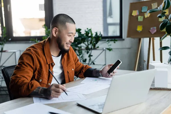 Smiling asian architect with mobile phone drawing blueprint near blurred laptop and house models — Stock Photo