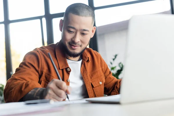 Smiling asian designer drawing near blurred laptop in architectural bureau — Stock Photo