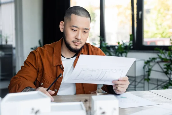 Bärtiger asiatischer Architekt arbeitet mit Bauplänen in der Nähe verschwommener Hausmodelle — Stockfoto