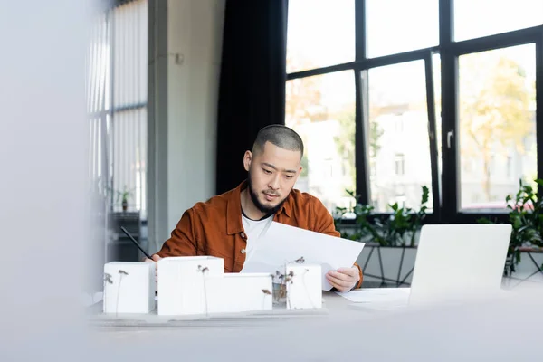 Asiatischer Architekt schaut sich Dokument an, während er in der Nähe verschwommener Laptop- und Hausmodelle arbeitet — Stockfoto