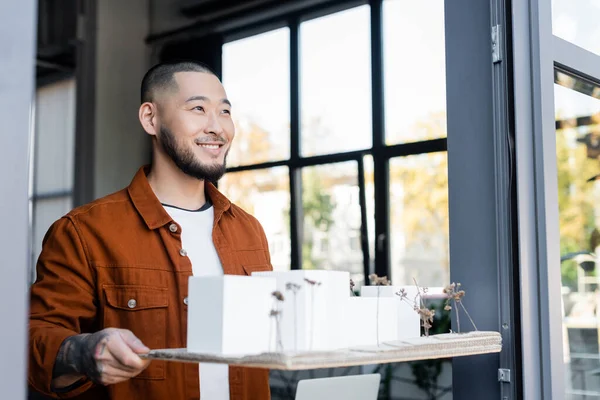 Glücklicher asiatischer Architekt schaut weg, während er Hausmodelle im Büro hält — Stockfoto