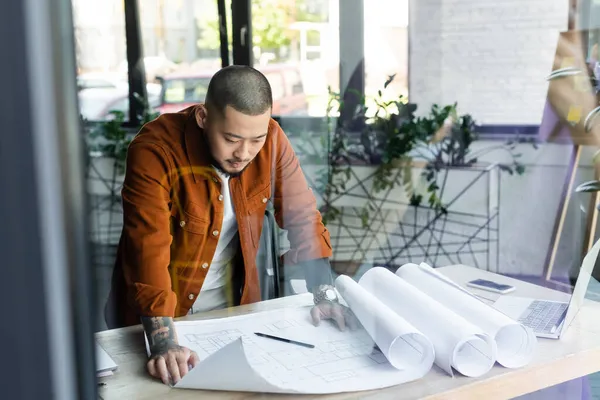 Asian architect looking at construction plan while standing at workplace on blurred foreground — Stock Photo