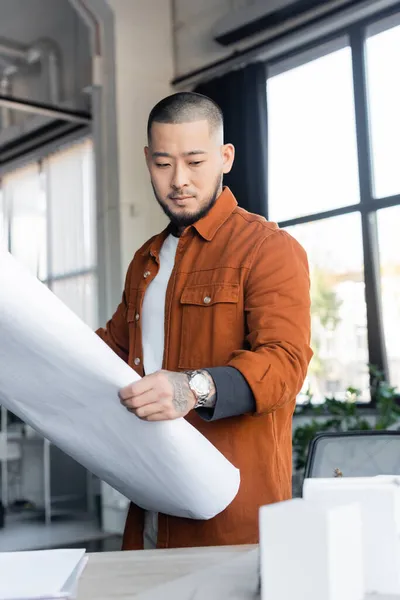 Asian architect looking at blurred house models while holding blueprint in office — Stock Photo