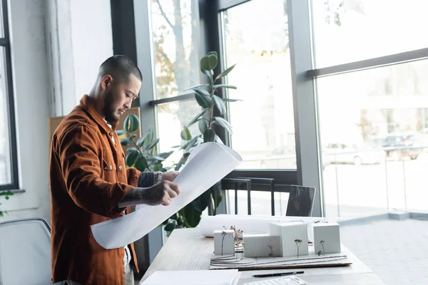 Asian architect holding blueprint while standing at workplace near house models — Stock Photo