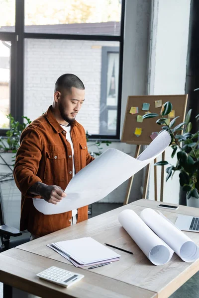 Asiatischer Ingenieur schaut sich Baupläne an, während er neben Papieren und Gadgets auf dem Schreibtisch steht — Stockfoto