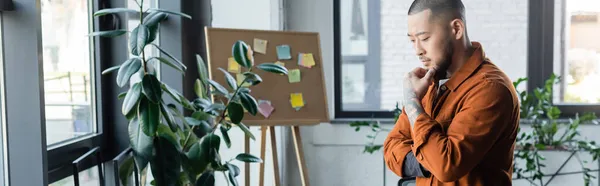 Pensive asian businessman thinking near blurred plants and note board in office, banner — Stock Photo