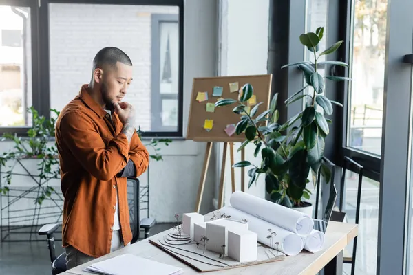 Pensativo asiático arquitecto mirando casa modelos en trabajo escritorio en oficina - foto de stock