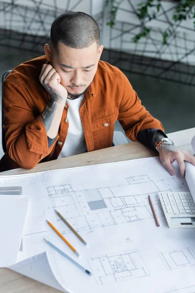 Nachdenklicher asiatischer Architekt betrachtet Bauplan, während er im Büro an einem Projekt arbeitet — Stockfoto