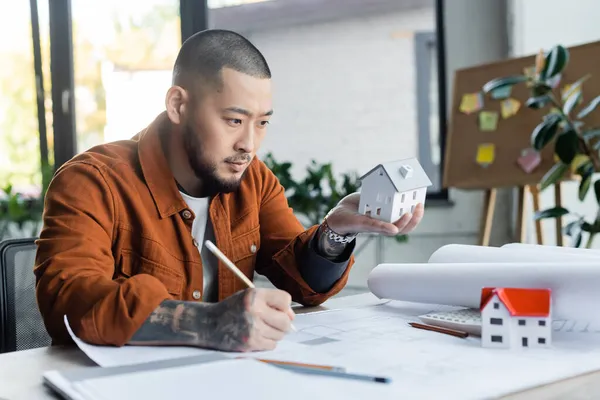 Asian architect holding house model while drawing blueprint in office — Stock Photo