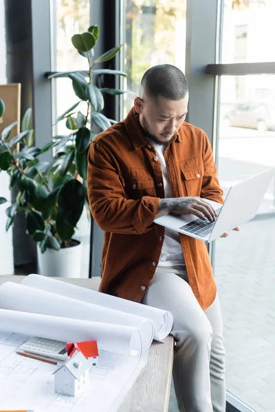 Konzentrierter asiatischer Architekt tippt am Laptop, während er am Schreibtisch neben Bauplänen und Hausmodellen sitzt — Stockfoto