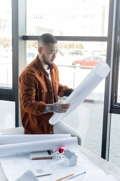 Asian architect holding blueprint while sitting on desk near papers and house models — Stock Photo