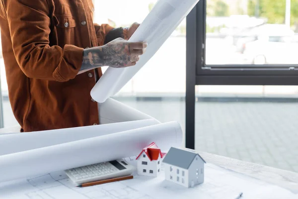 Ausgeschnittene Ansicht eines tätowierten Architekten mit Blaupause, während er am Schreibtisch in der Nähe von Taschenrechnern und Hausmodellen sitzt — Stockfoto