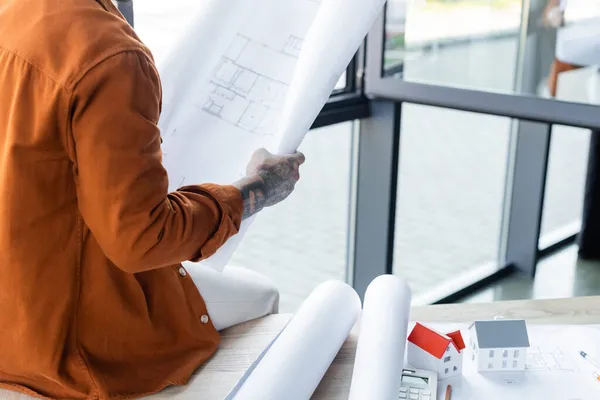 Geschnittene Ansicht des Architekten mit Bauplan, während er auf dem Schreibtisch in der Nähe von Bauplänen und Hausmodellen sitzt — Stockfoto