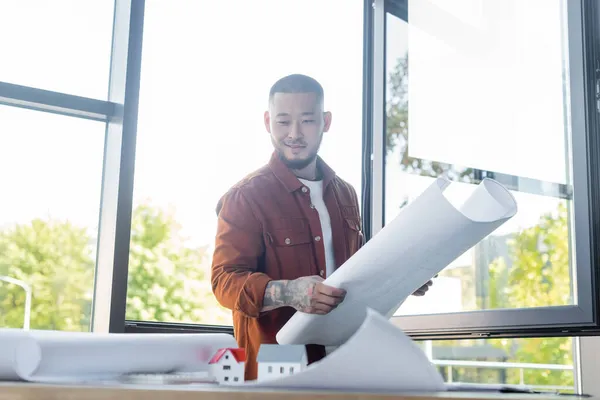 Smiling asian architect looking at blurred house models while holding blueprint in office — Stock Photo