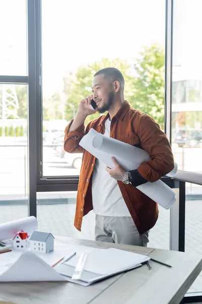 Glücklicher asiatischer Architekt mit gerollten Bauplänen, der im Büro mit dem Smartphone spricht — Stockfoto