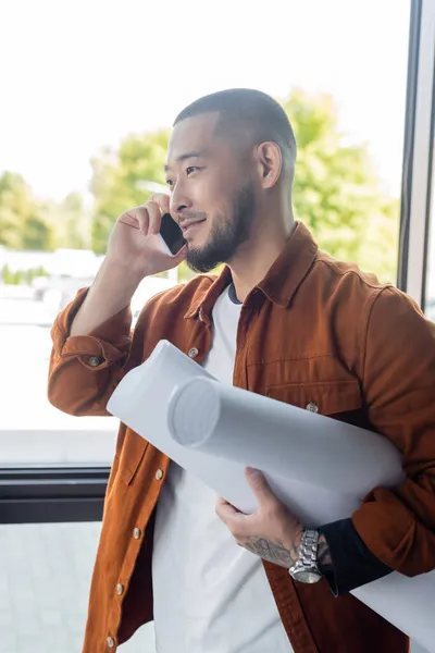 Bearded asian architect with rolled blueprints smiling during phone conversation in office — Stock Photo