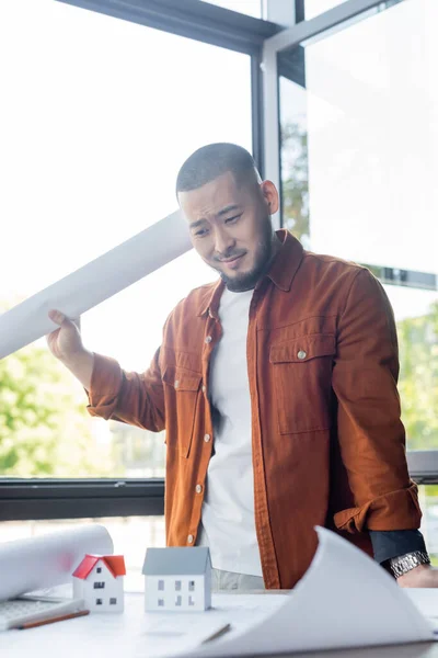 Displeased asian architect holding blueprint while standing near house models and papers on work desk — Stock Photo