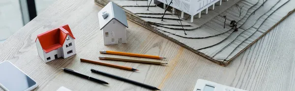 High angle view of pencils, house models and smartphone on wooden desk, banner — Stock Photo