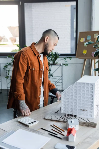 Татуированный азиатский архитектор, стоящий за рабочим столом и смотрящий на модель дома в офисе — стоковое фото