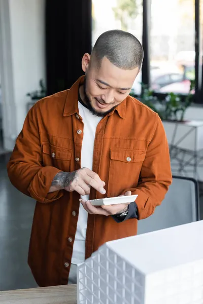 Tatuado asiático arquitecto sonriendo mientras usando calculadora cerca borrosa casa modelo - foto de stock
