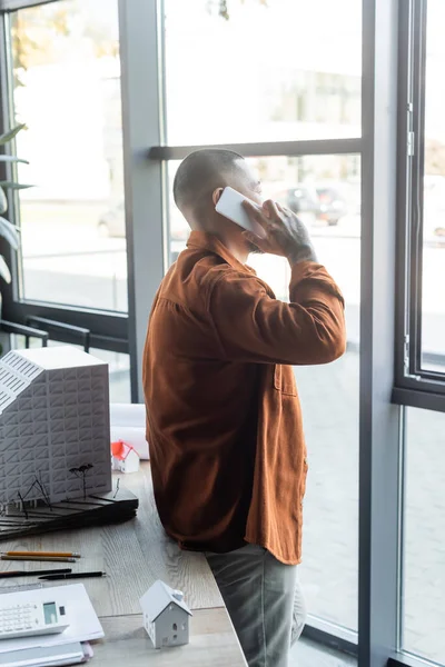 Asiatischer Architekt telefoniert mit dem Handy, während er neben Fenstern und Hausmodellen auf dem Schreibtisch steht — Stockfoto
