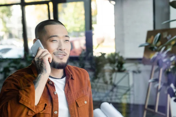 Веселый азиатский бизнесмен смотрит в сторону, разговаривая по мобильному телефону в офисе — стоковое фото