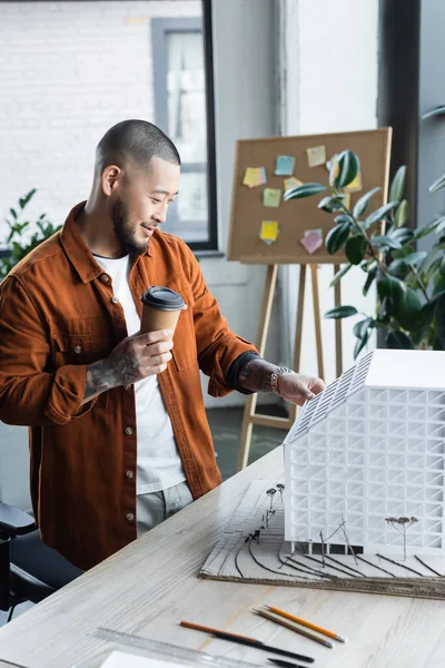 Азіатський архітектор, дивлячись на модель будинку, стоячи на робочому столі з кавою, щоб піти — стокове фото