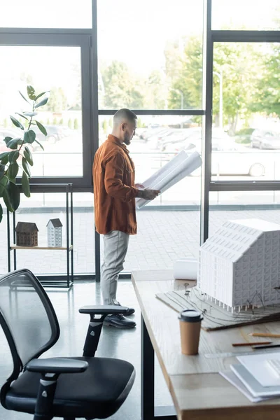 Arquitecto asiático con plano de pie cerca de ventanas y escritorio con modelo de casa en la oficina - foto de stock