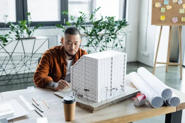 Asiático arquitecto sentado cerca casa modelo mientras trabajo en arquitectura proyecto en oficina - foto de stock