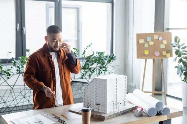 Arquitecto asiático sosteniendo lápiz mientras habla en teléfono inteligente cerca de modelos de casa en el escritorio - foto de stock