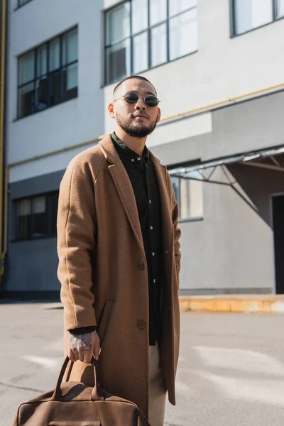 Élégant asiatique homme en lunettes de soleil et beige manteau debout près flou bâtiment à l'extérieur — Photo de stock