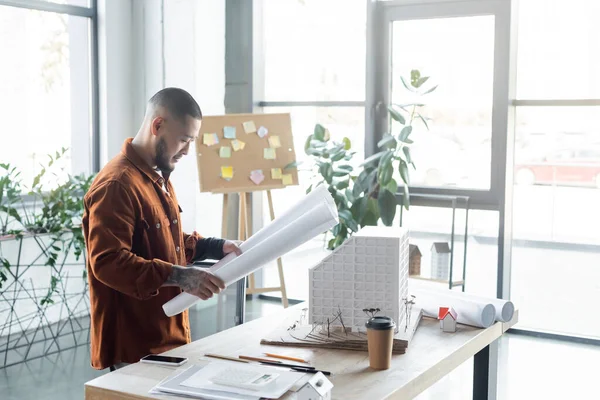 Arquiteto asiático olhando para blueprint enquanto pé perto de casa modelo e smartphone na mesa — Fotografia de Stock