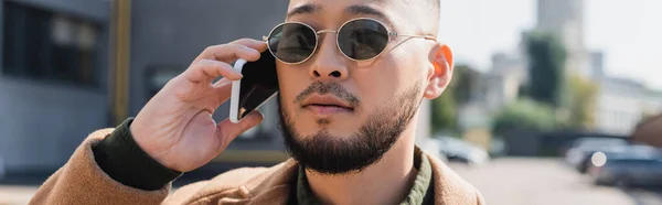 Asian man in stylish sunglasses talking on smartphone outdoors, banner — Stock Photo