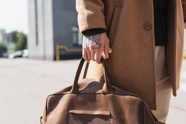 Vista parcial del hombre tatuado en abrigo beige que sostiene el maletín de cuero al aire libre — Stock Photo