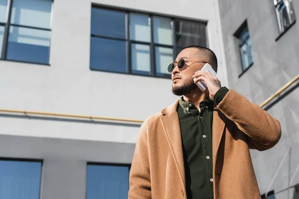 Low angle view of asian man in sunglasses and coat calling on smartphone in city — Stock Photo