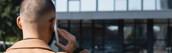 Back view of tattooed man talking on mobile phone near blurred building, banner — Stock Photo