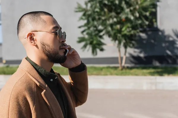 Side view of bearded asian man in sunglasses talking on smartphone on city street — Stock Photo