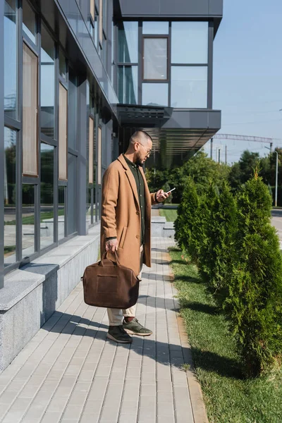 Full length view of asian man with leather briefcase using mobile phone near building with glass facade — Stock Photo