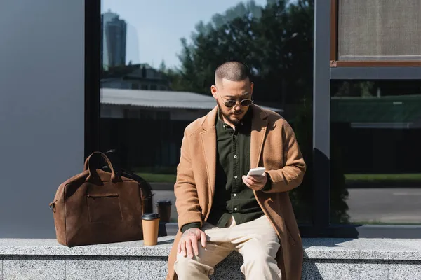 Stylish asian man using smartphone on border near takeaway drink and leather briefcase — Stock Photo